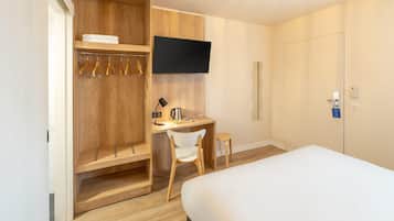 Habitación de hotel con cama, escritorio con silla, televisor montado en la pared y armario con ropa colgada.