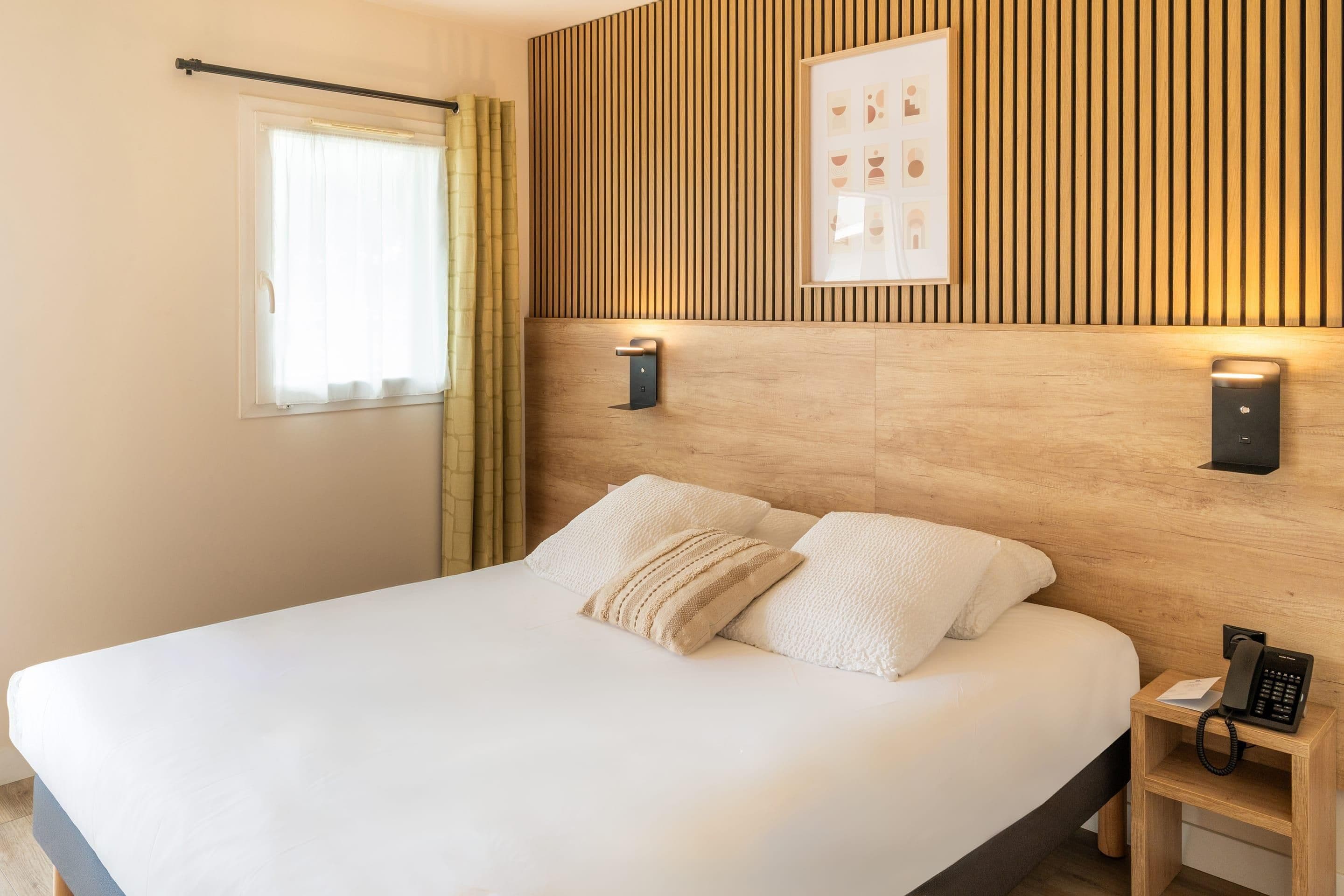 A hotel room with a large bed, a wooden headboard, a bedside table with a phone, and a window with curtains.