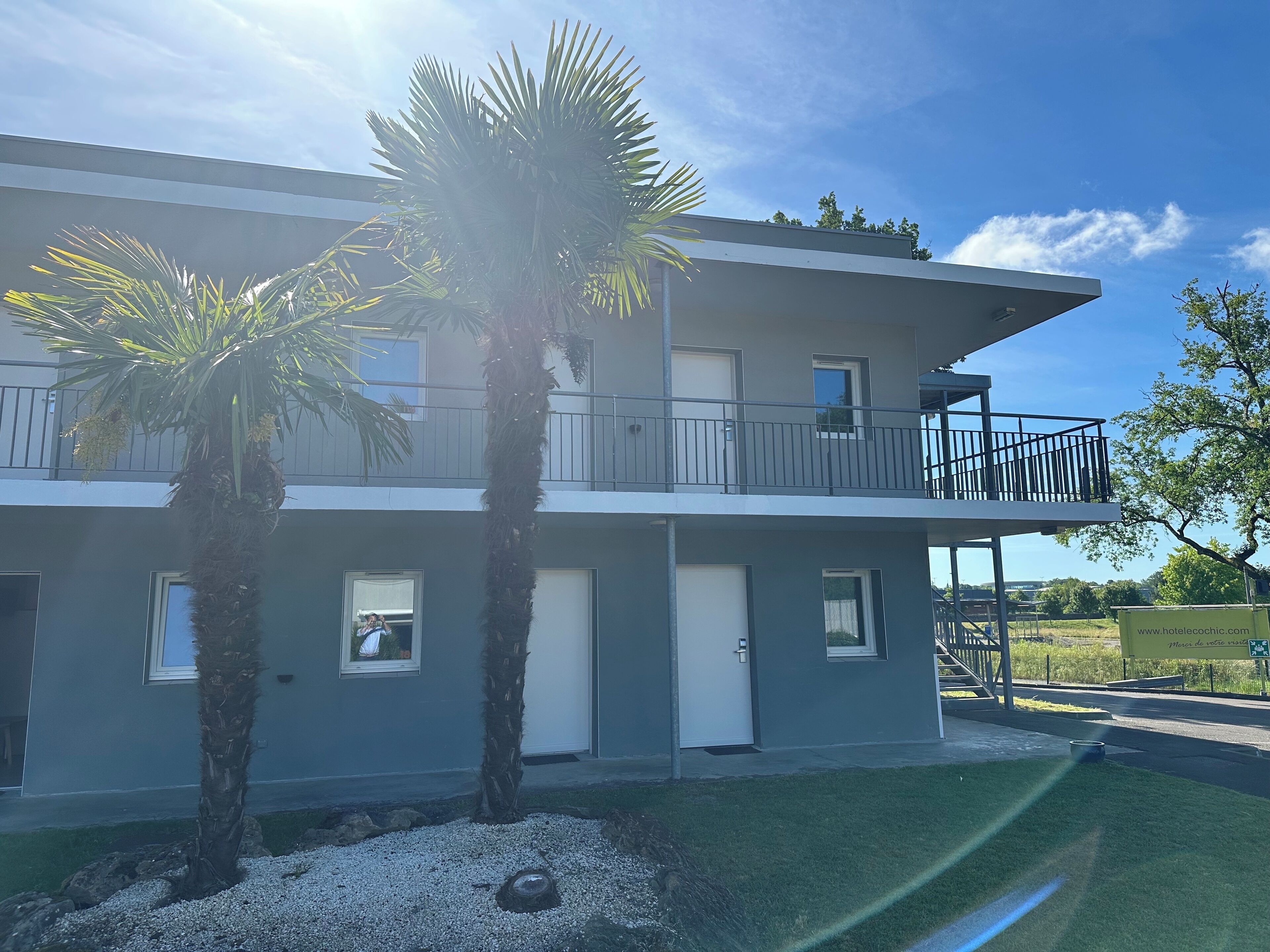 A two-story building with a balcony, palm trees, and a clear sky.