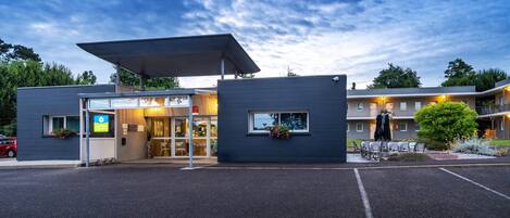 A modern hotel with a covered entrance, outdoor seating, and a parking lot.