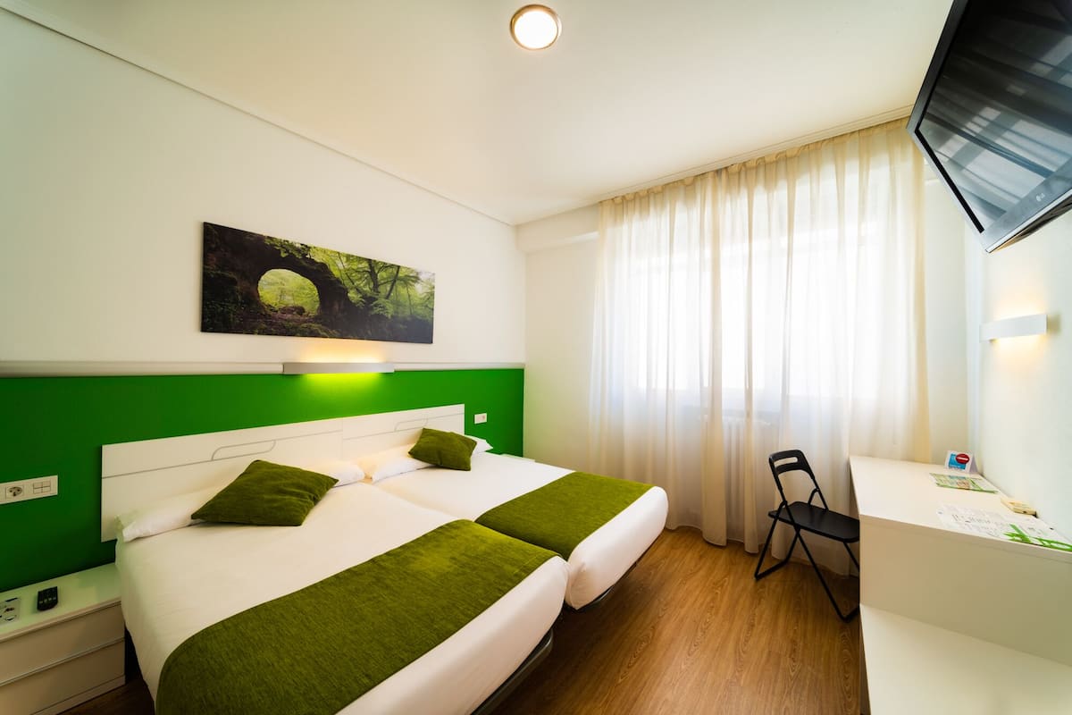 Hotellihuone, jossa on yksi seinä maalattu vihreällä värillä, valkoisilla lakanoilla varustettu sänky, työpöytä tuolilla ja maalaus, jossa on kuvattuna metsämaisema.