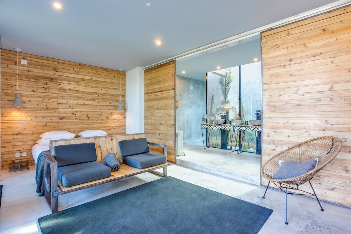Ein modernes Zimmer mit einer Holzwand, einem Sofa, einem Sessel und einem Essbereich, der durch eine Glastür sichtbar ist.