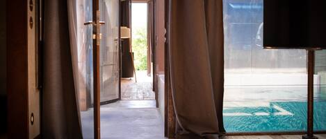 Une porte ouverte donne sur une terrasse en bois avec une piscine, des rideaux sont disposés de part et d’autre, et un téléviseur se trouve sur le côté droit.