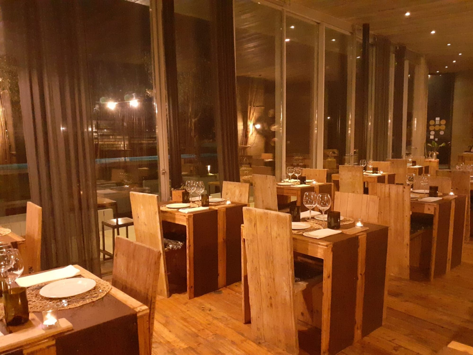 A restaurant with wooden tables and chairs, large windows, and a warm interior lighting.