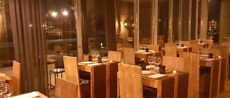A restaurant with wooden tables and chairs, large windows, and a warm interior lighting.