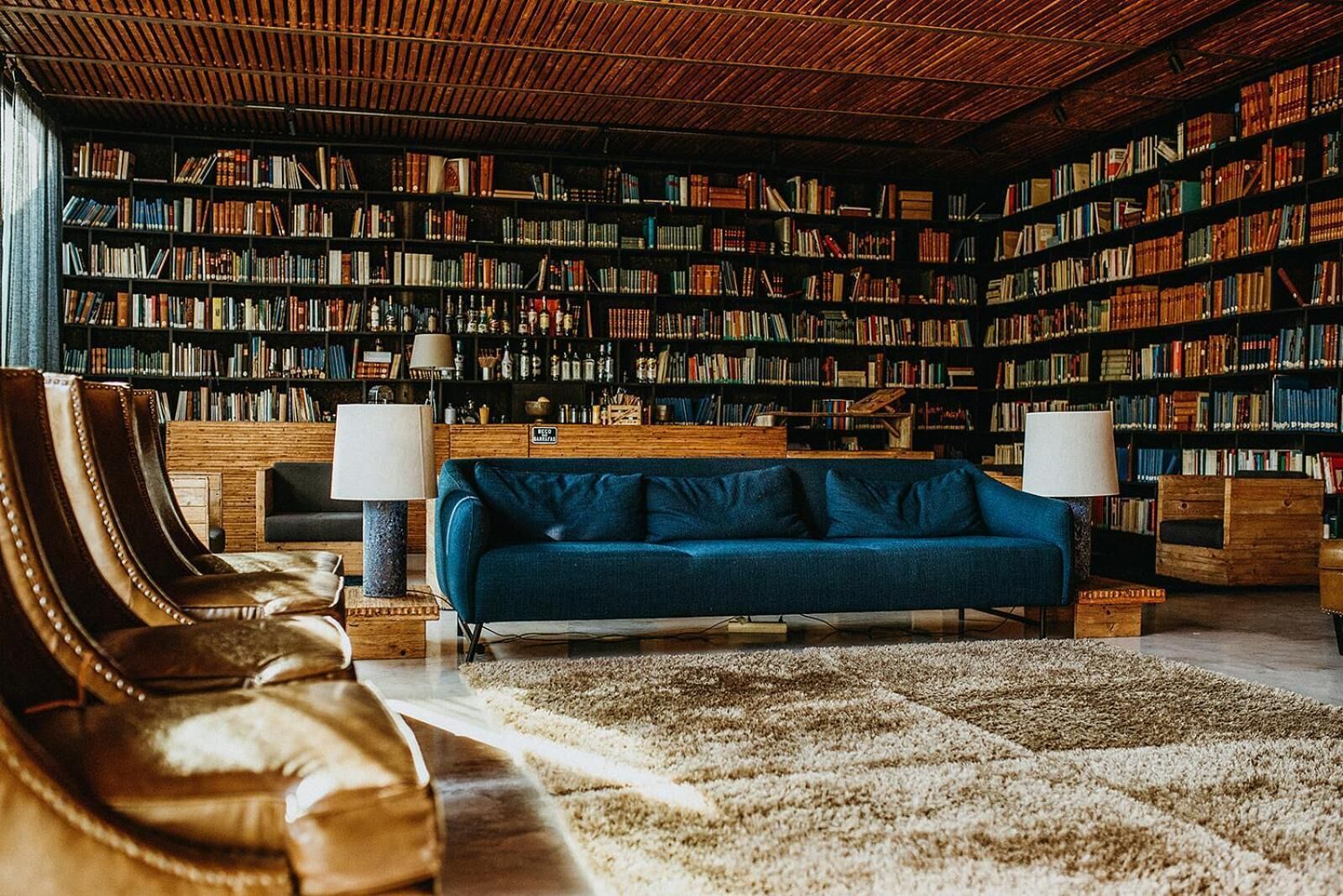 A library with bookshelves, a blue sofa, leather chairs, and two lamps.