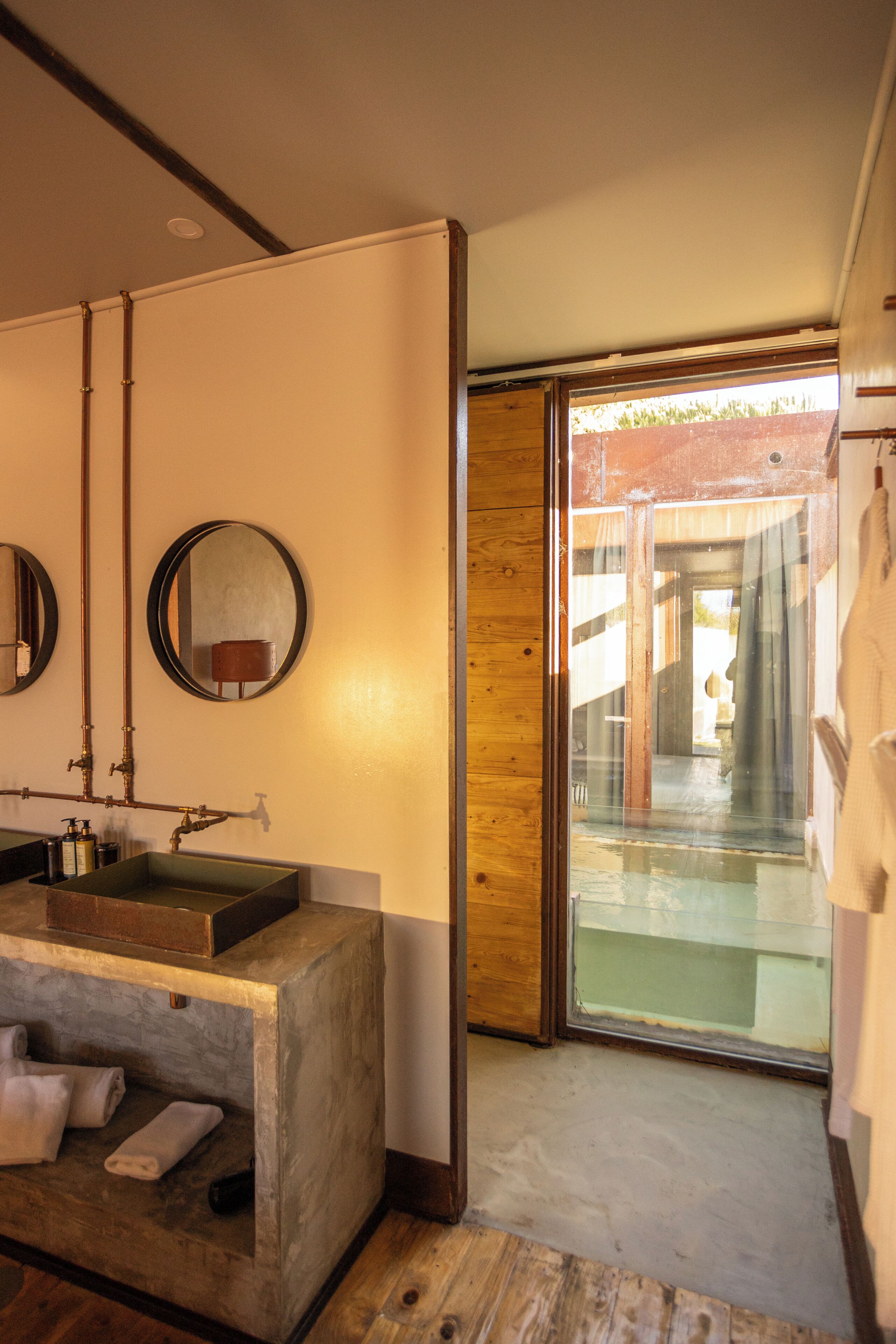 Une salle de bain avec un meuble-lavabo en bois, deux miroirs ronds et une porte en verre donnant sur un balcon.