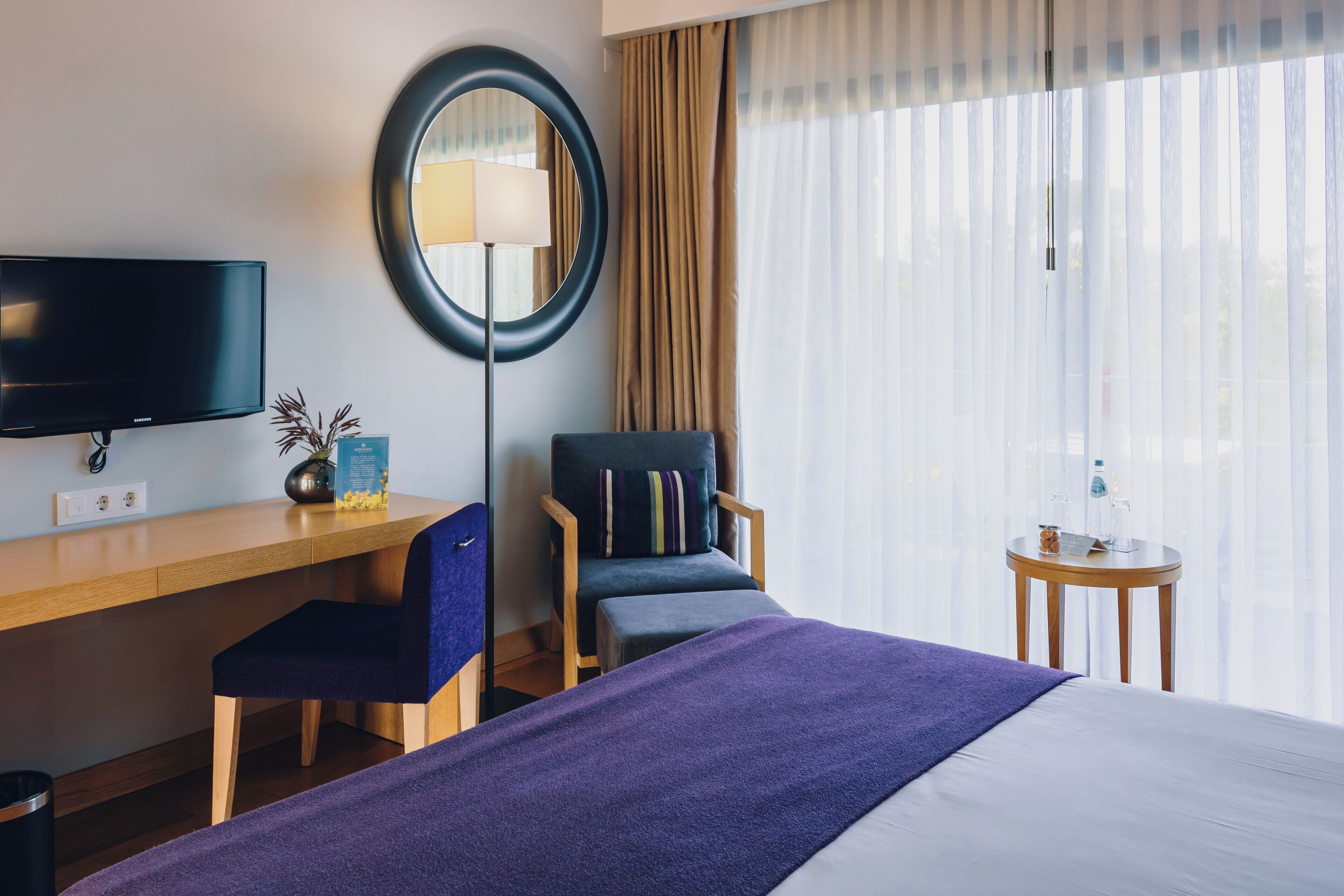 Habitación de hotel con cama, escritorio con silla, televisor, lámpara, mesita y ventana con cortinas.