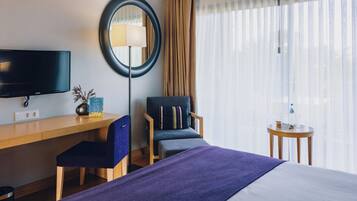 Habitación de hotel con cama, escritorio con silla, televisor, lámpara, mesita y ventana con cortinas.