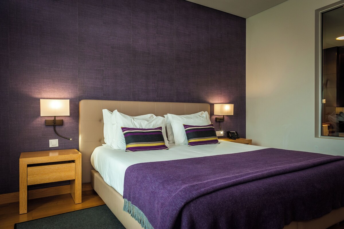 Una habitación de hotel con una cama, dos mesitas de noche, una lámpara de noche, una manta púrpura y dos lámparas fijadas a la pared.
