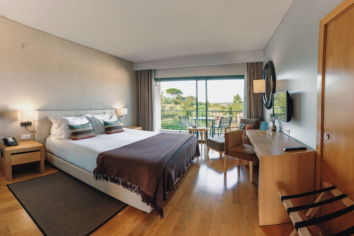 A modern hotel room with a large bed, a desk, and a balcony with a view of the outdoors.