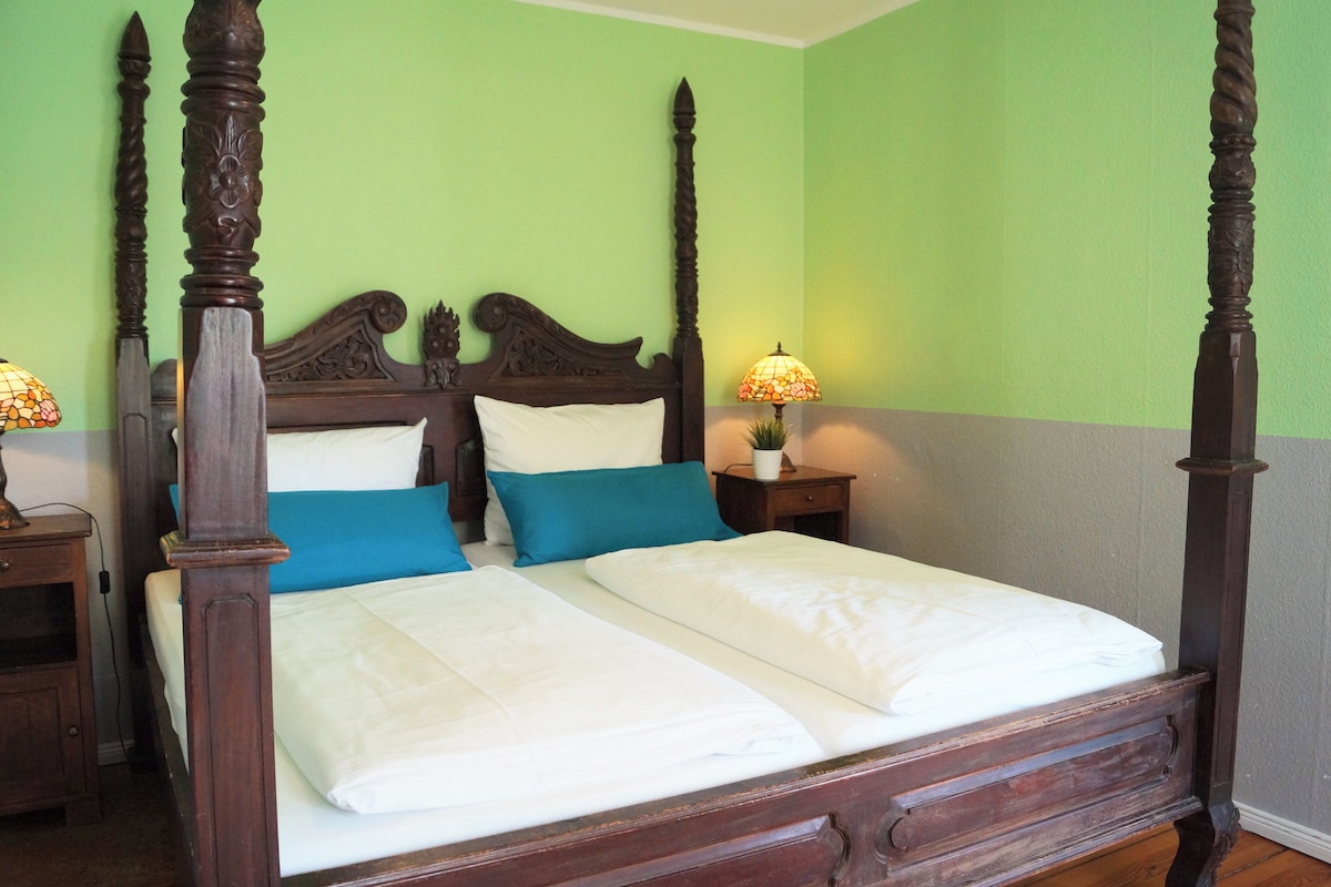 Una cama con dosel, ropa de cama blanca, dos almohadas azules y una mesita de noche con lámpara.