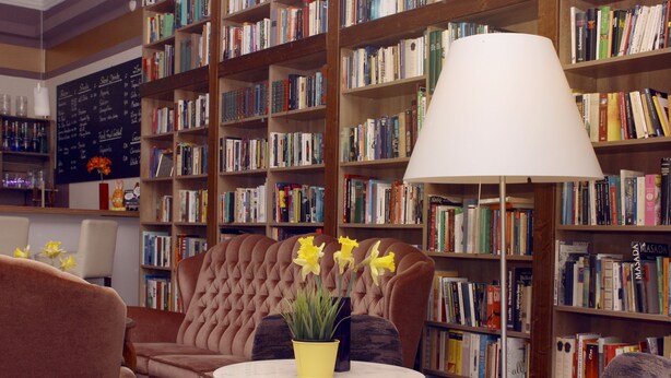 A library with bookshelves, a comfortable armchair, a table with a yellow vase, and a large lamp. - Grand Hostel Berlin Classic (Berlin)