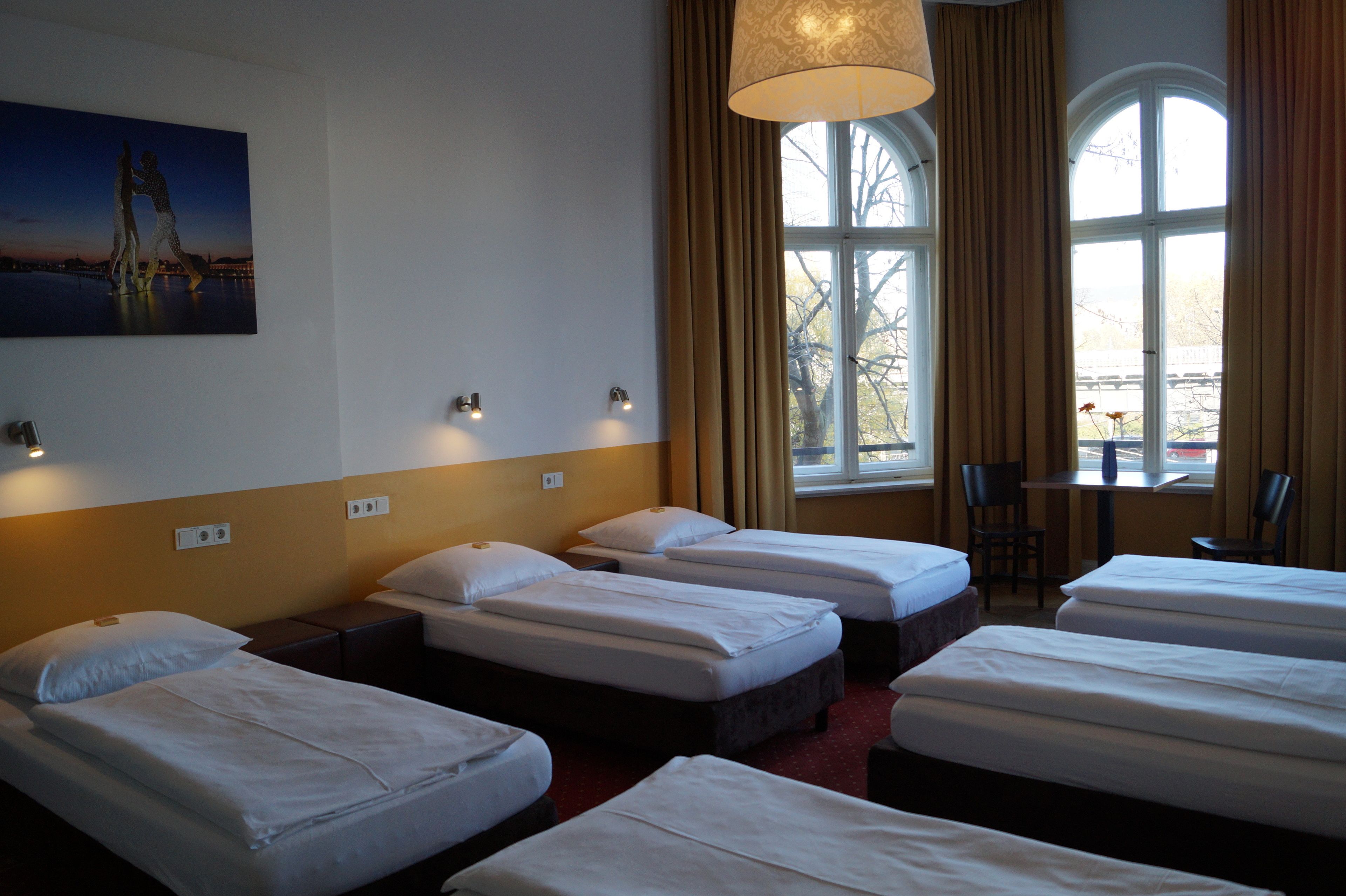 Habitación de hotel con cuatro camas individuales, un cuadro en la pared, una mesa con sillas y una ventana con cortinas.