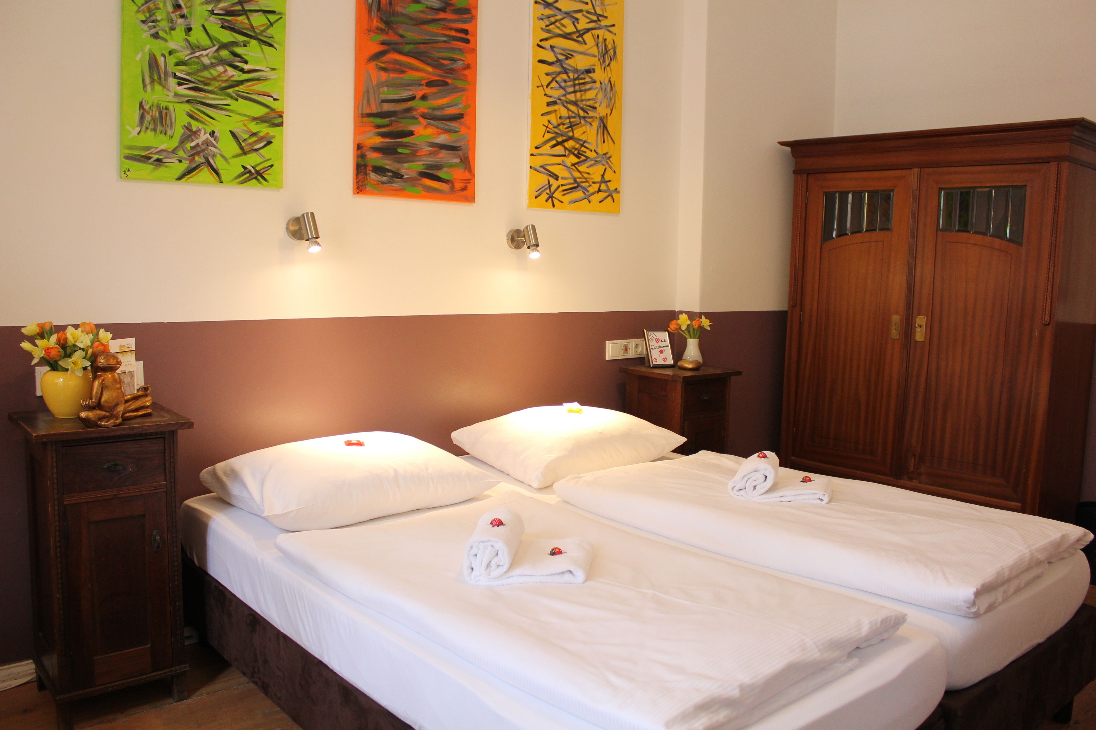 A hotel room with two beds, a wooden wardrobe, and three abstract paintings on the wall.