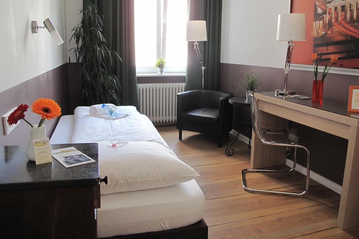 A hotel room with a bed, a desk, a chair, a radiator, and a window with curtains.