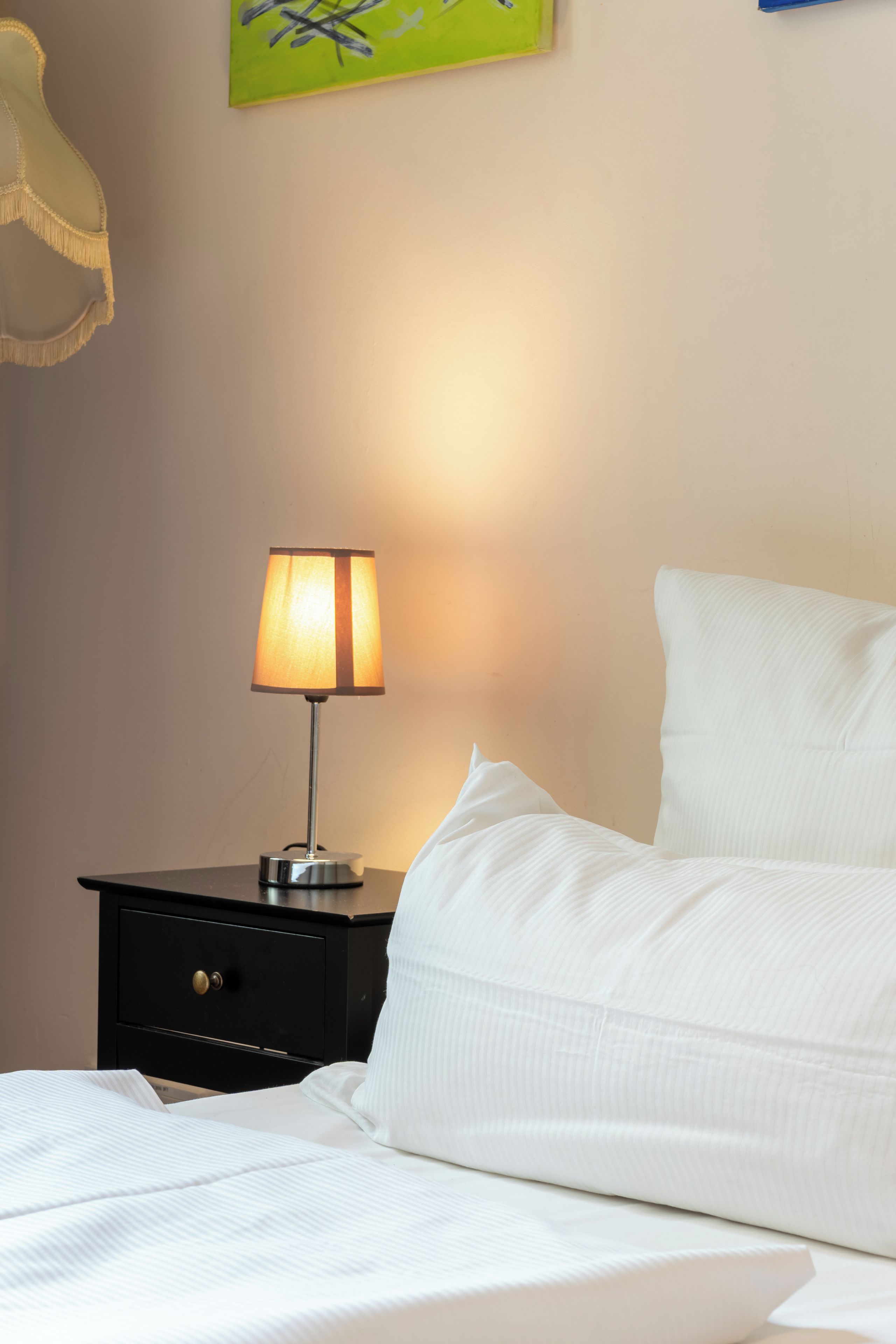 A neatly made bed with white bedding, a bedside lamp, and a small black nightstand with a drawer.
