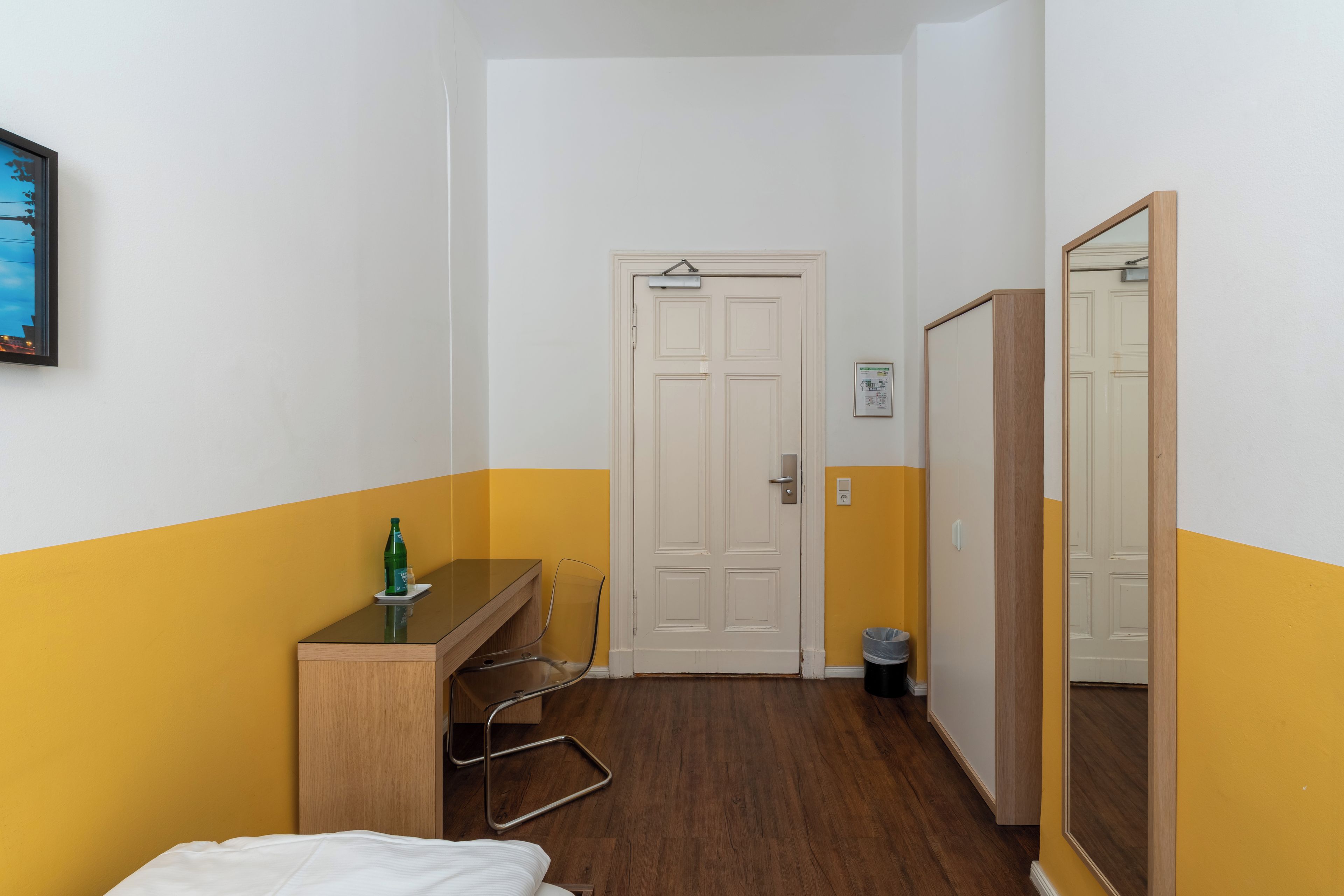 Habitación de hotel con cama, escritorio, silla, espejo, puerta y basurero.
