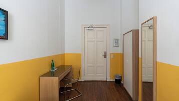 Habitación de hotel con cama, escritorio, silla, espejo, puerta y basurero.