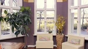 A room with a wooden table, two sofas with colorful cushions, a large potted plant, and a window with a view of a street and buildings. - Grand Hostel Berlin Classic (Berlin)