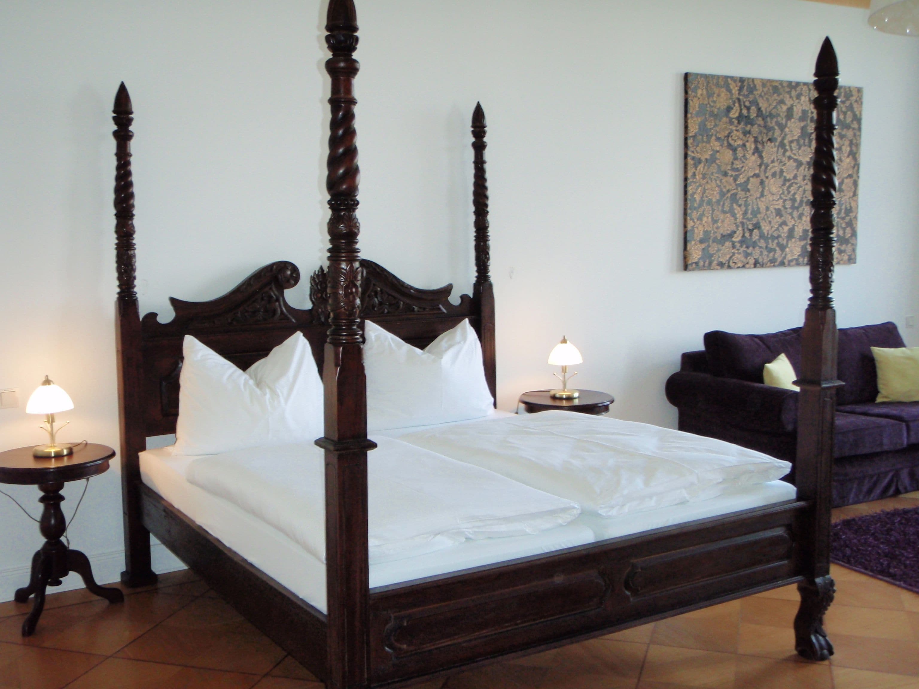 A four-poster bed with white linens, two bedside tables with lamps, a purple sofa, and a floral wall art piece.