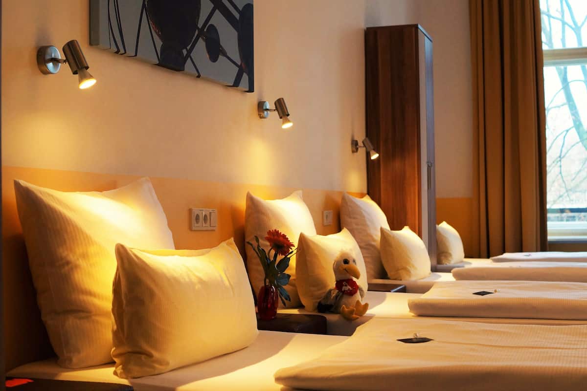 A hotel room with multiple beds, each with white bedding and pillows. There are wall-mounted lamps providing light, and a painting on the wall.