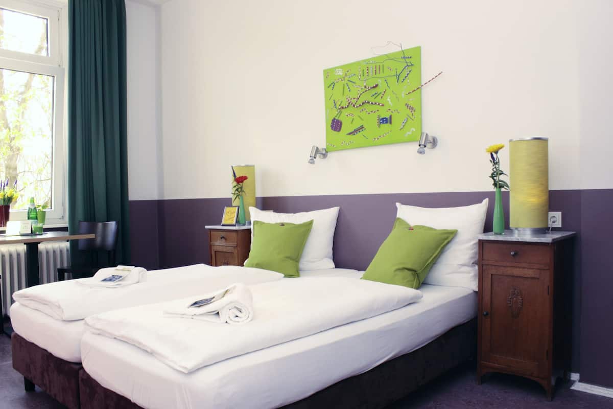A double bed with green and white pillows, a small table with a vase and a book, and a green wall art piece.