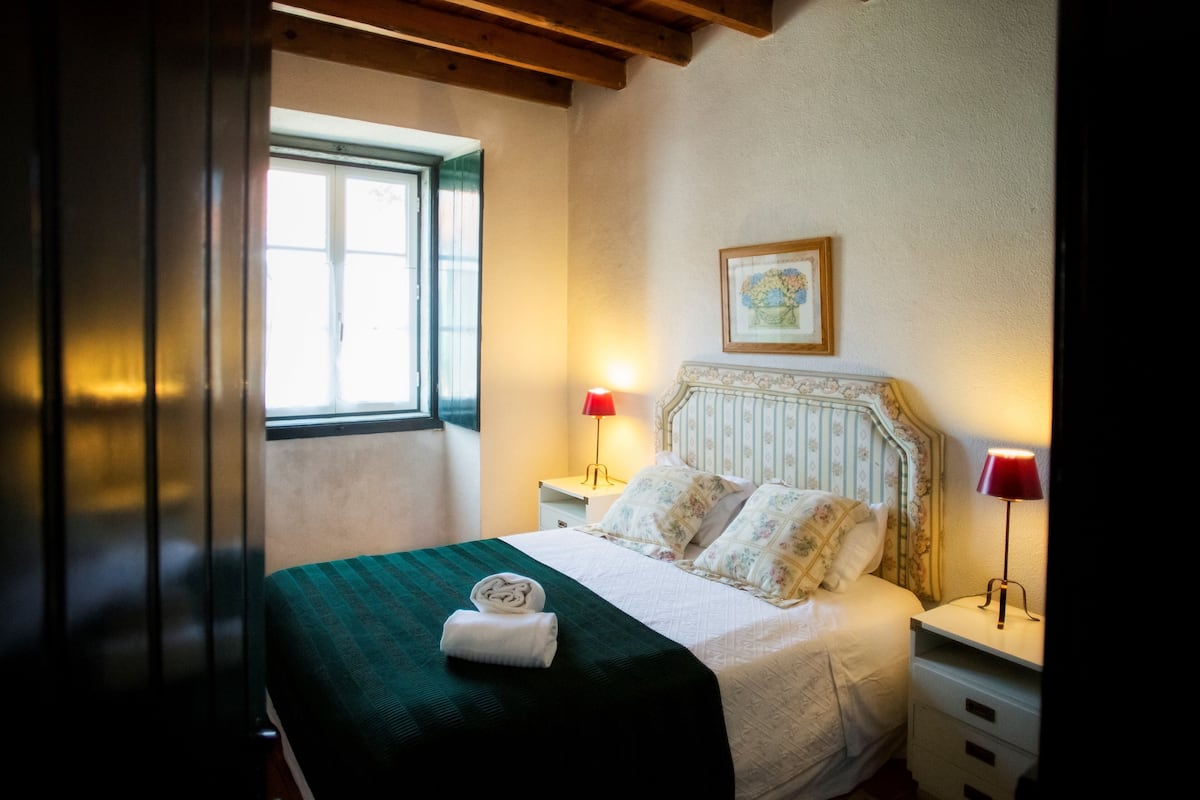 A neatly made bed with a green bedspread and two pillows, two bedside lamps, a framed picture on the wall, and a window with curtains.