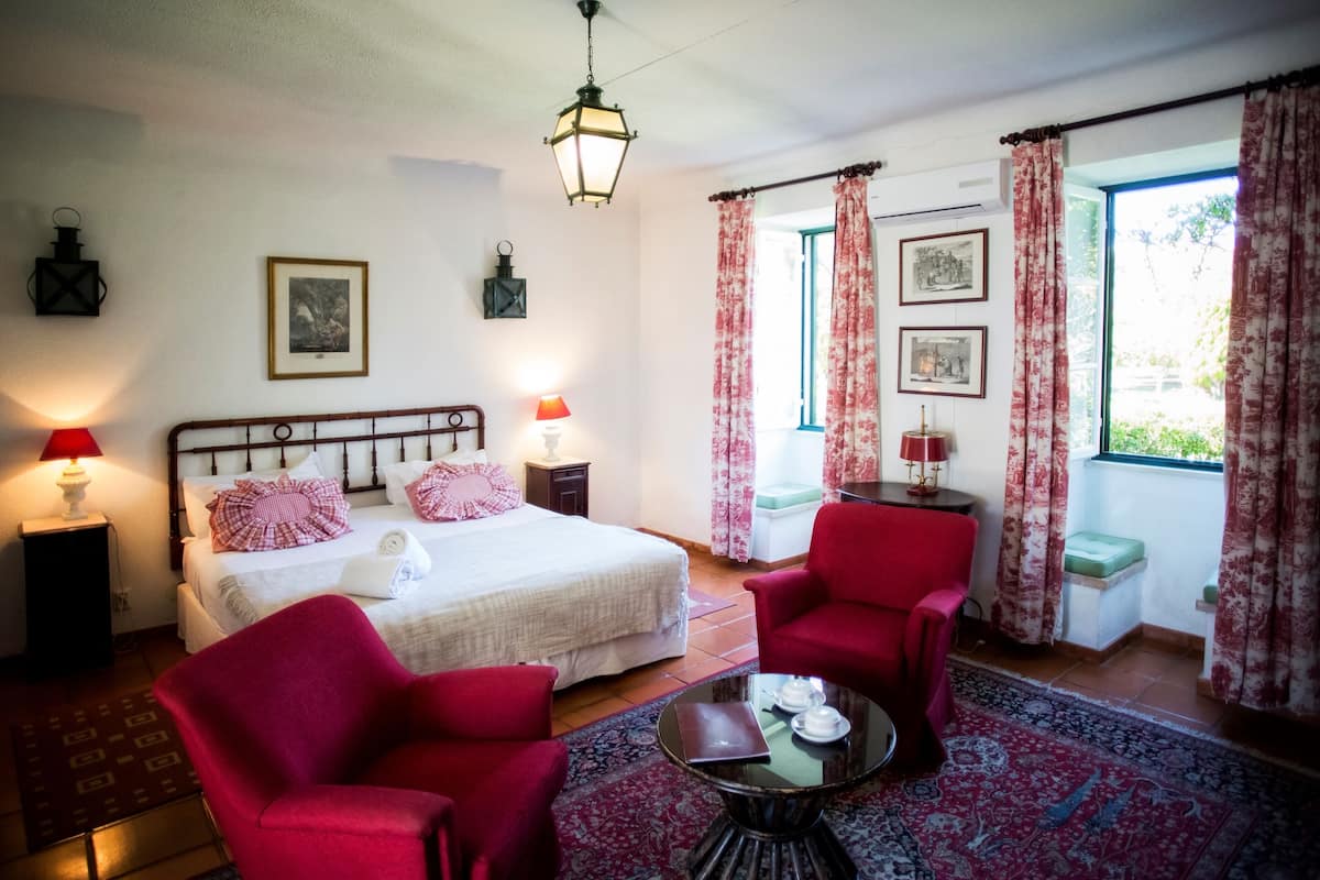 A hotel room with a bed, two red chairs, a small table with a laptop and teapot, a nightstand with a lamp, and a window with curtains.