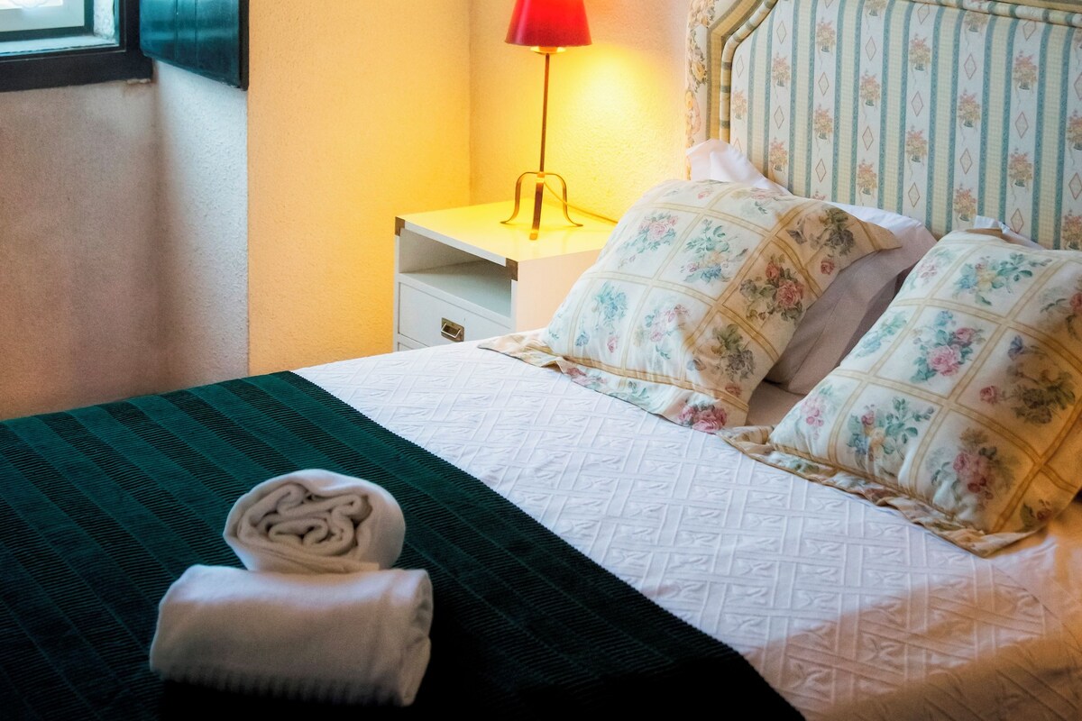 A neatly made bed with floral pillows, a red lamp on a bedside table, and a window with blinds.