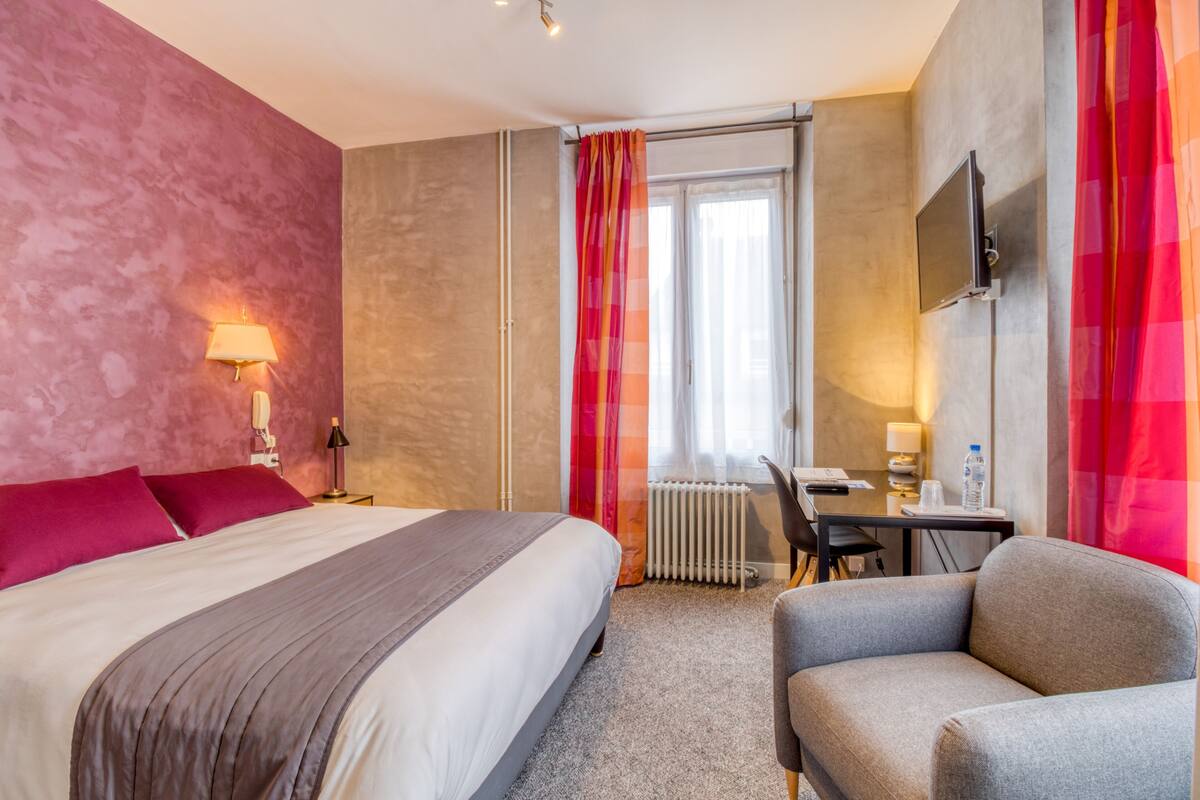 Une chambre d’hôtel avec un lit, un bureau, une chaise, une télévision et une fenêtre avec des rideaux.