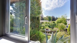 Une fenêtre donnant sur un jardin où l’on peut admirer différentes plantes et un abreuvoir à oiseaux.
