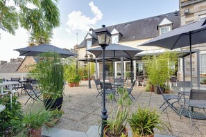 Ein Terrassenbereich im Freien mit Tischen und Stühlen, umgeben von Pflanzgefäßen und Sonnenschirmen.