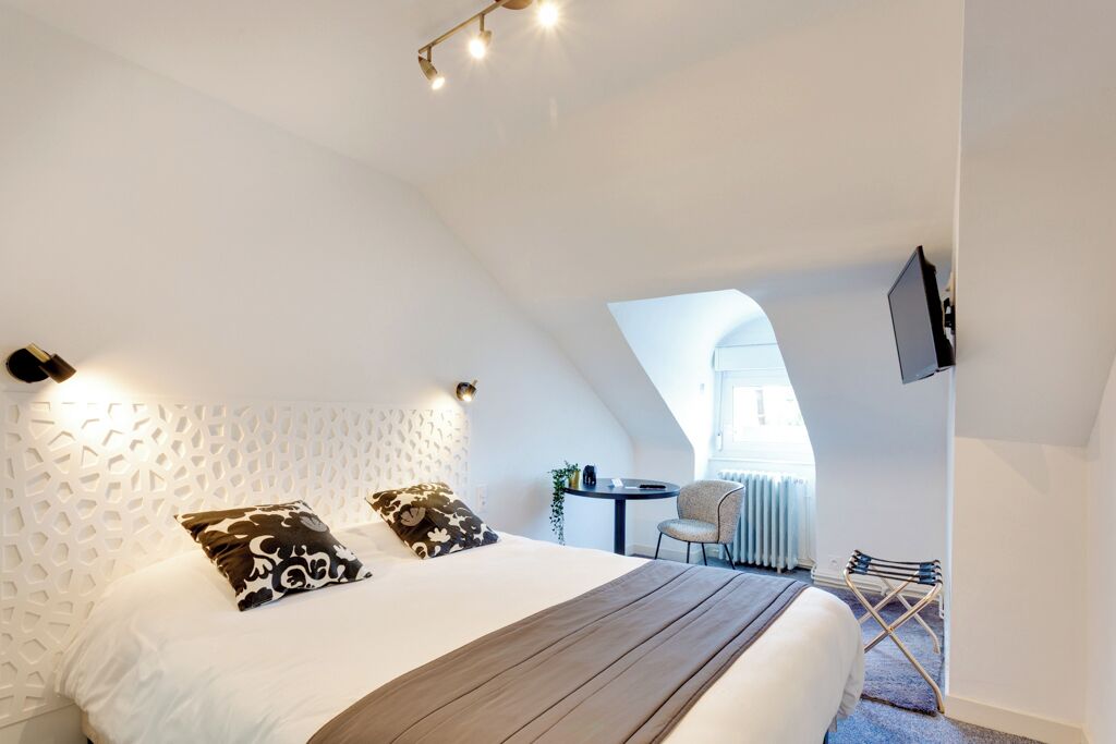 Un dormitorio moderno con techo inclinado, una cama con almohadas estampadas, una mesita con una silla y un televisor montado en la pared.