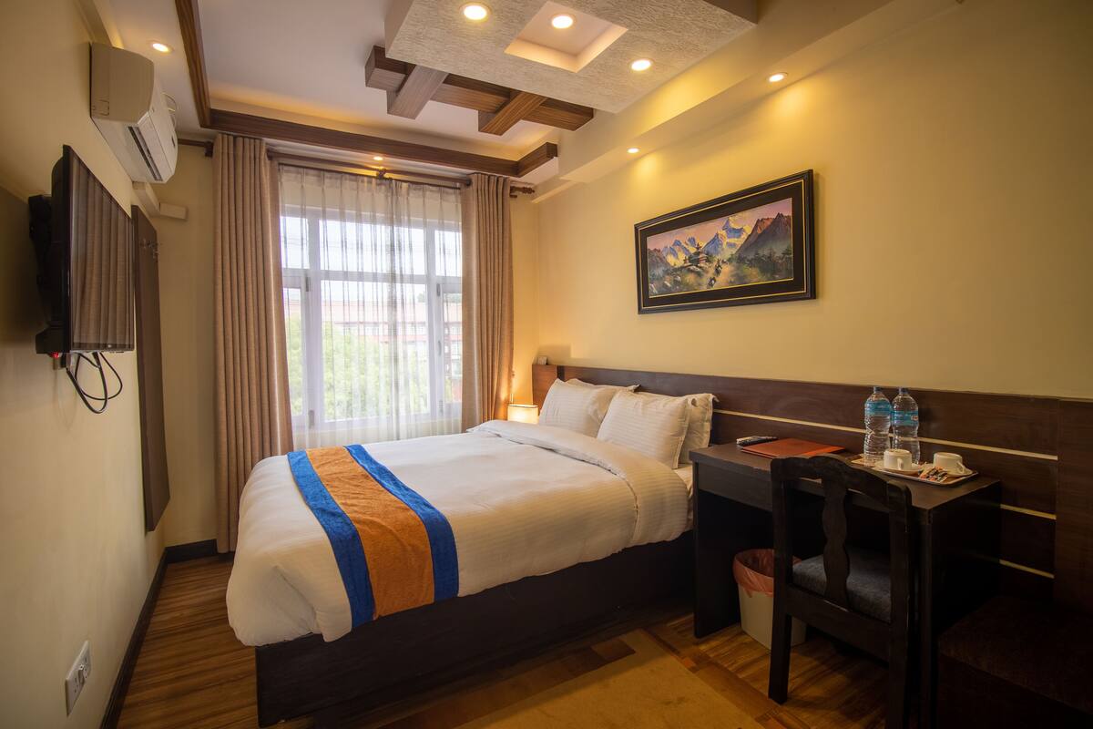 A hotel room with a bed, a desk, a chair, a painting, and a window with curtains.