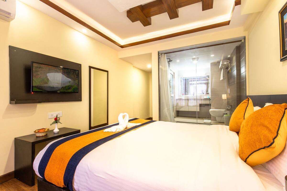 A hotel room with a bed, a TV, a mirror, a bedside table with a vase and fruit basket, and a sliding glass door to the bathroom.