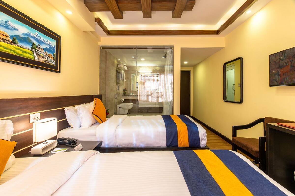 A hotel room with two beds, a desk, a chair, a bathroom visible through a glass door, and a painting on the wall.