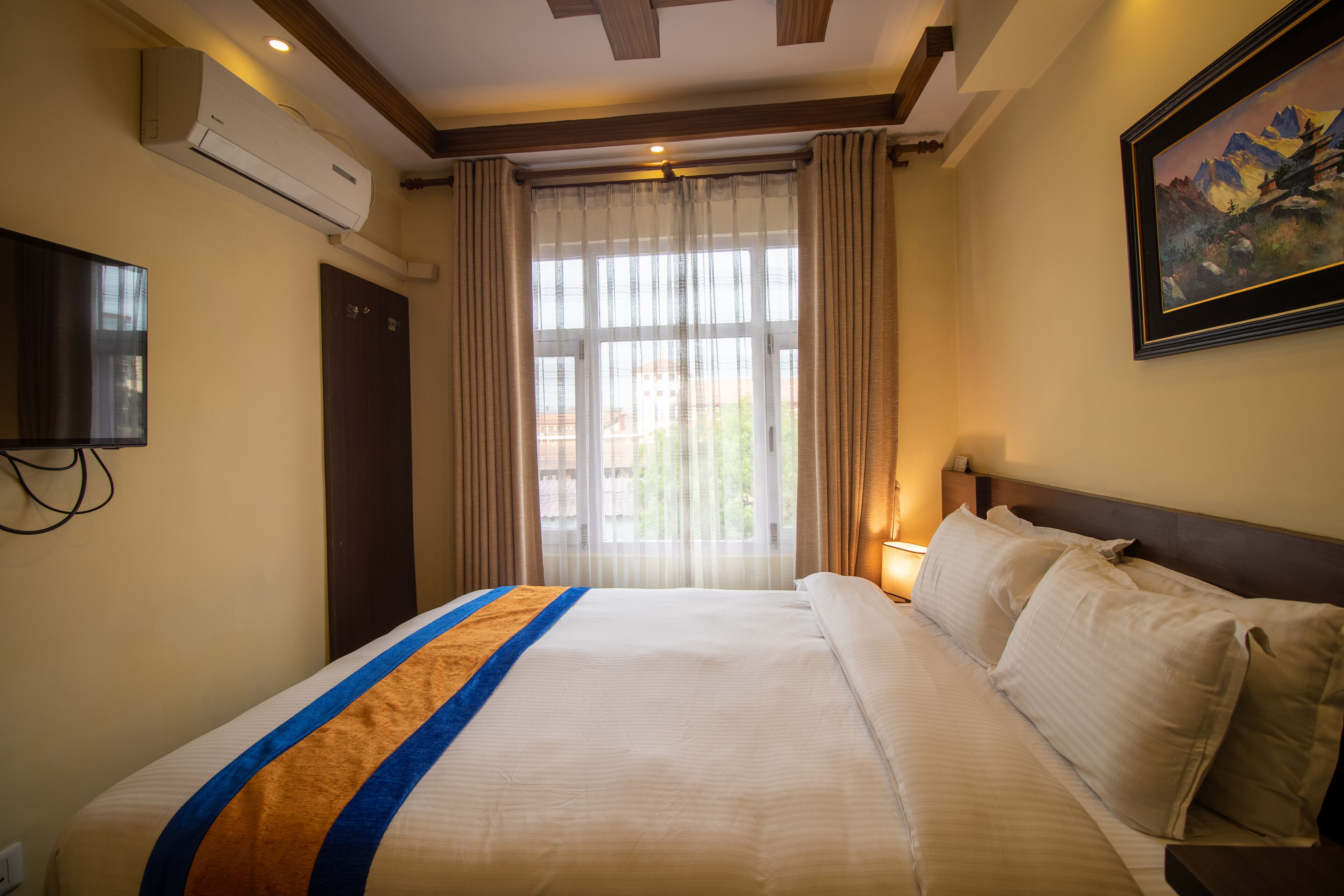 A hotel room with a large bed, a flat-screen TV, an air conditioning unit, a painting on the wall, and a window with curtains.