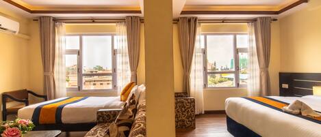 Habitación de hotel con dos camas, un sofá, una mesita con un florero y ventanas amplias con cortinas.