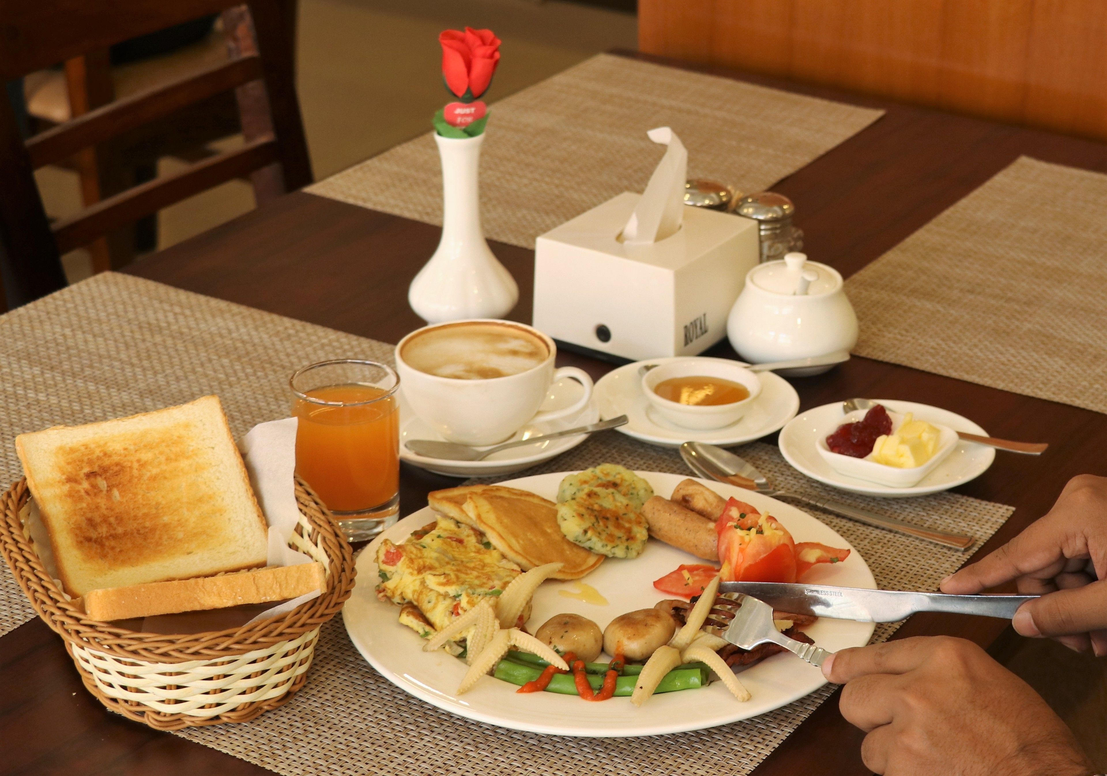Un desayuno con tostadas, huevos, salchichas y verduras en un plato, una taza de café y un jarrón con una rosa.