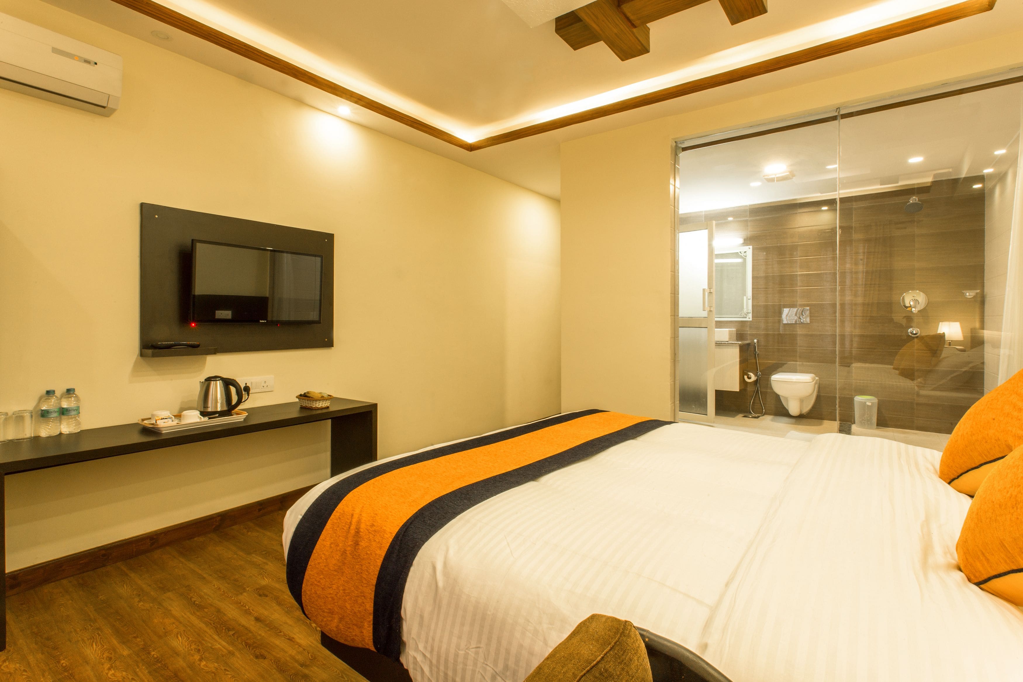 A hotel room with a bed, bedside table, TV, and a bathroom with a glass shower.