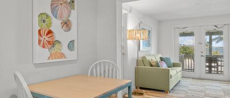 Et spiseområde med et bord og stole, en grøn sofa med puder, en gulvlampe og et stort maleri af sømuslinger på væggen.