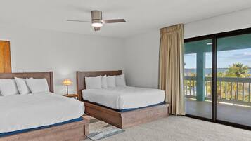 Quarto de hotel com duas camas, um ventilador de teto, mesa de cabeceira com abajur e vista para o mar através de amplas portas deslizantes.