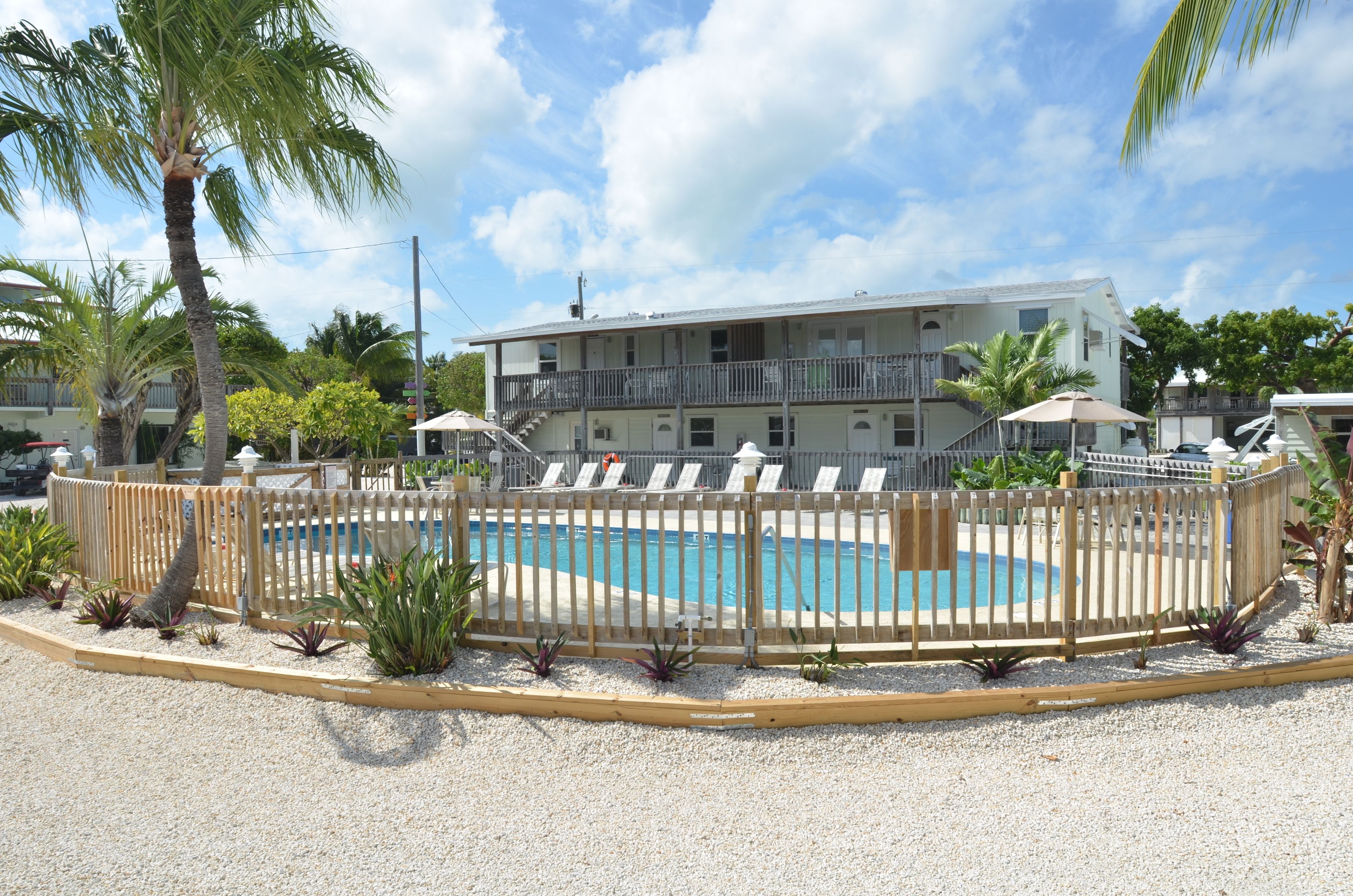 A pool area with a wooden fence, lounge chairs, and a building with balconies in the background.