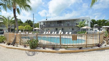 A pool area with a wooden fence, lounge chairs, and a building with balconies in the background.