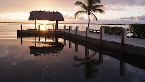 Vista al atardecer con una estructura de techo de paja y una palmera junto a un cuerpo de agua tranquilo.