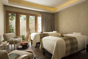 A hotel room with two beds, a sitting area with a chair and table, a small table with a bowl and a book, and a view of the outdoors through a sliding door.