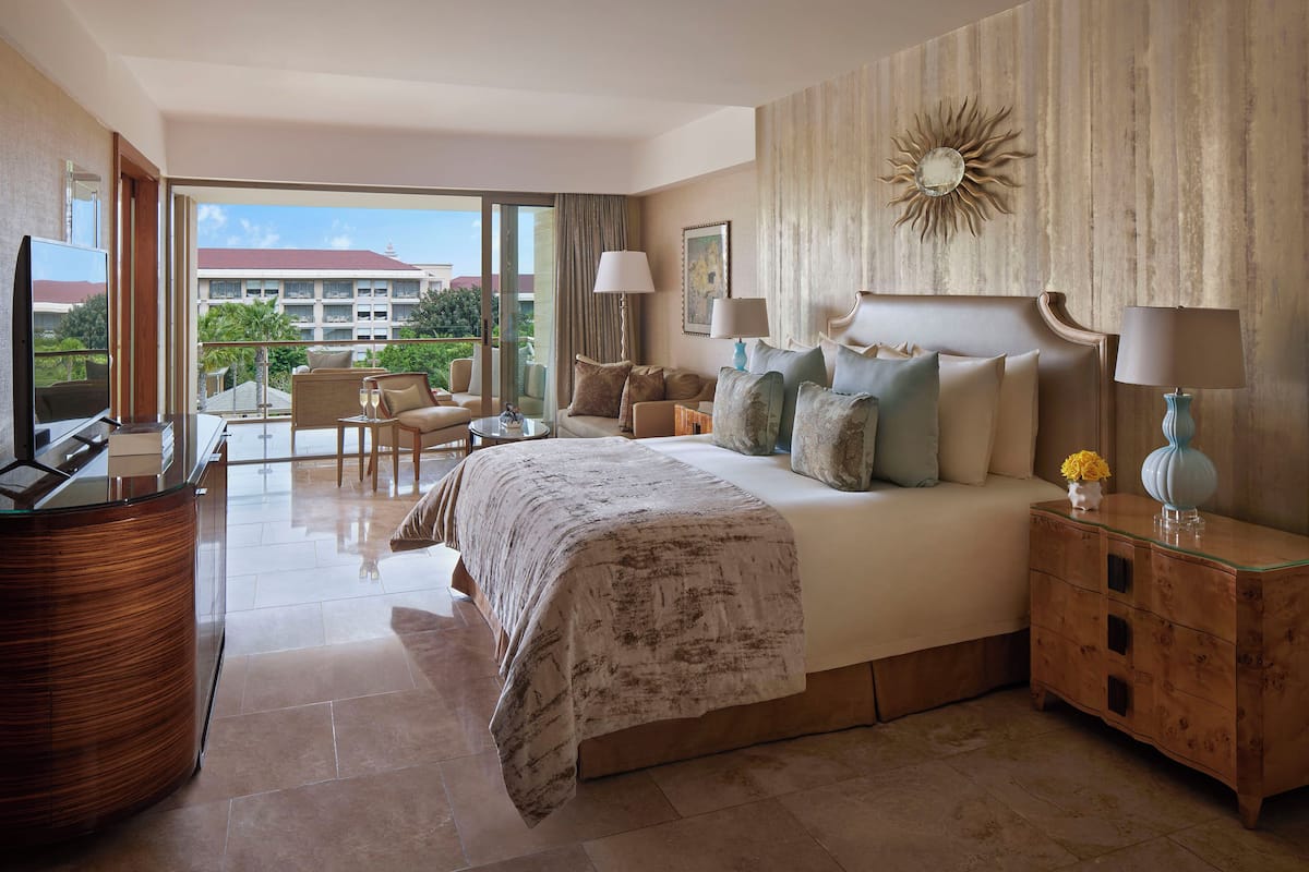 Una habitación de hotel con una cama grande, una zona de estar con sofá y sillones, una mesa de centro de vidrio, un televisor y una mesita de noche de madera con lámparas.