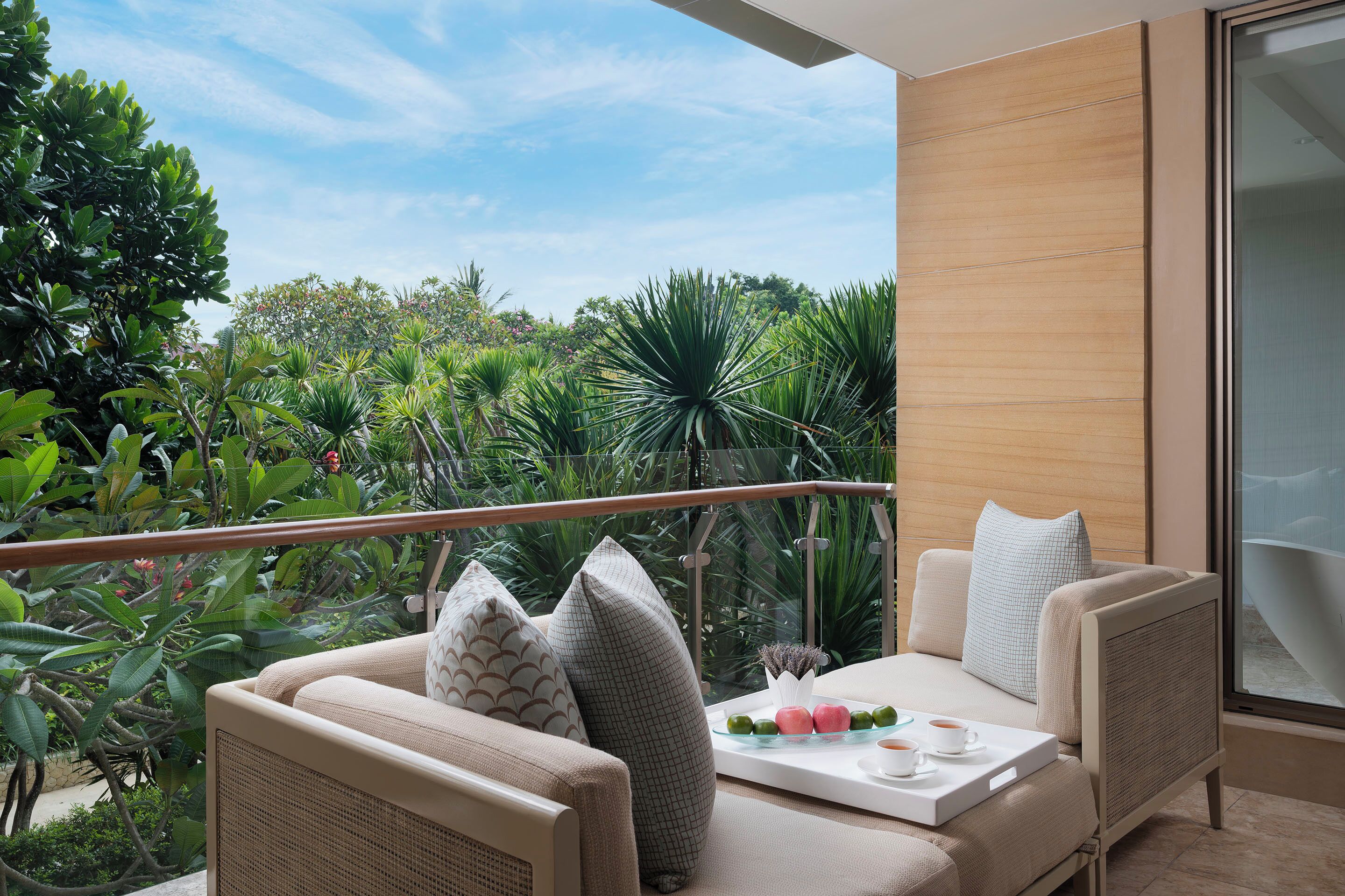 A balcony with wicker furniture, a glass table with a tray of items, and a view of lush greenery.