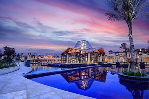 A modern hotel with a large pool, a dome-shaped glass structure, and palm trees. - Mulia Resort (Nusa Dua)